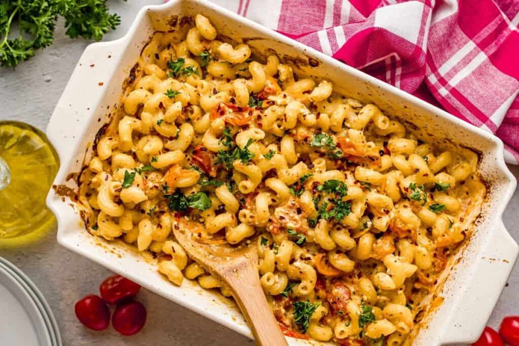 Baked macaroni and cheese in a white casserole dish, garnished with parsley and sun-dried tomatoes&mdash;a comforting pasta dinner&mdash;with a wooden spoon and a red checkered cloth nearby.