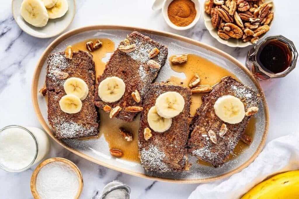 Four slices of French toast topped with banana slices, pecans, and powdered sugar on a plate&mdash;surrounded by syrup, pecans, and cinnamon&mdash;for one of the tastiest breakfast ideas.