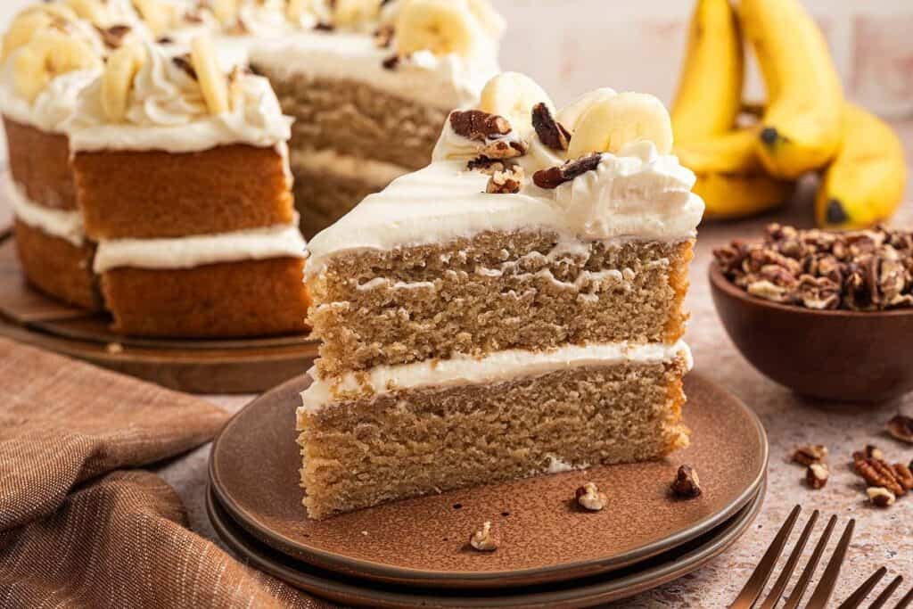 A slice of two-layer banana cake with white frosting, topped with banana slices and nuts, on a plate. Whole cake, bananas, and nuts visible in the background&mdash;perfect for those seeking easy desserts to impress.