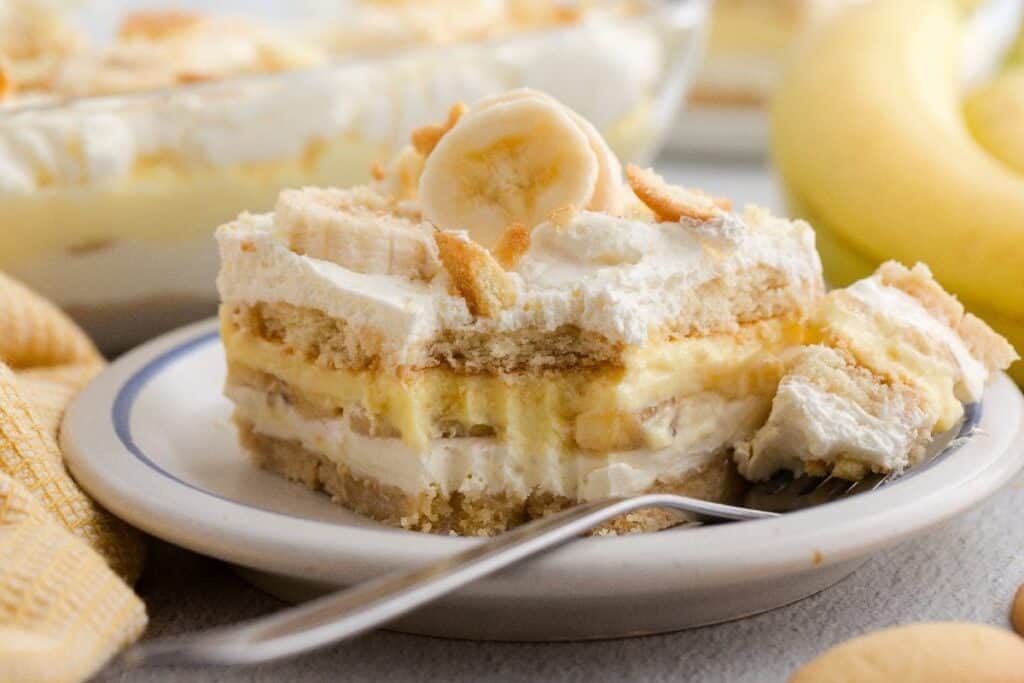 A slice of banana pudding dessert, one of the best Easy Desserts, with layers of cookies, banana slices, and creamy filling, topped with whipped cream and banana, served on a plate with a fork.