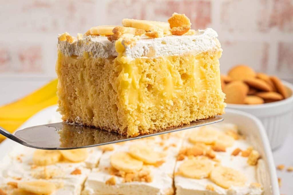 A close-up of a square slice of banana pudding poke cake with whipped topping and banana slices, held on a spatula above a baking dish&mdash;an irresistible treat perfect for fans of easy desserts.