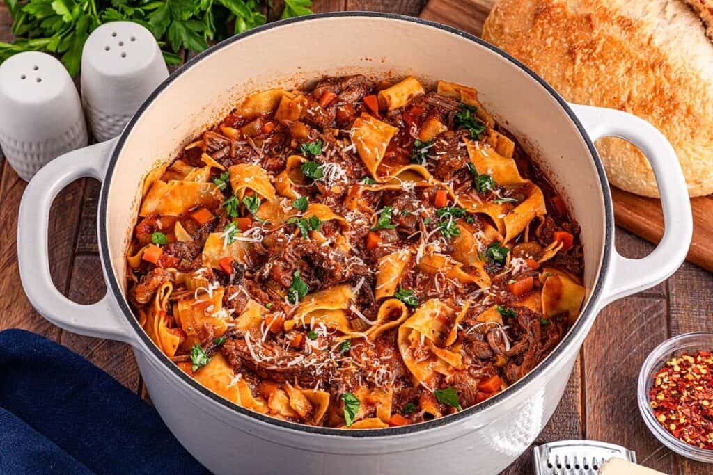A white Dutch oven filled with pasta dinner in a rich beef and tomato sauce, garnished with parsley and grated cheese, sits on a wooden table next to bread and seasonings.