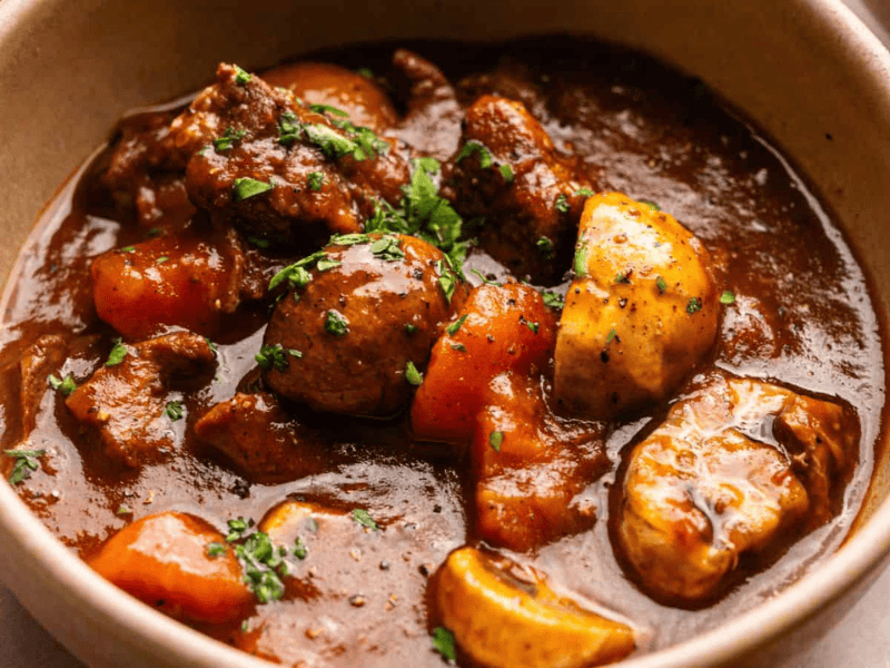 A bowl of beef stew with chunks of beef, carrots, potatoes, and mushrooms in a thick brown broth, garnished with chopped herbs.