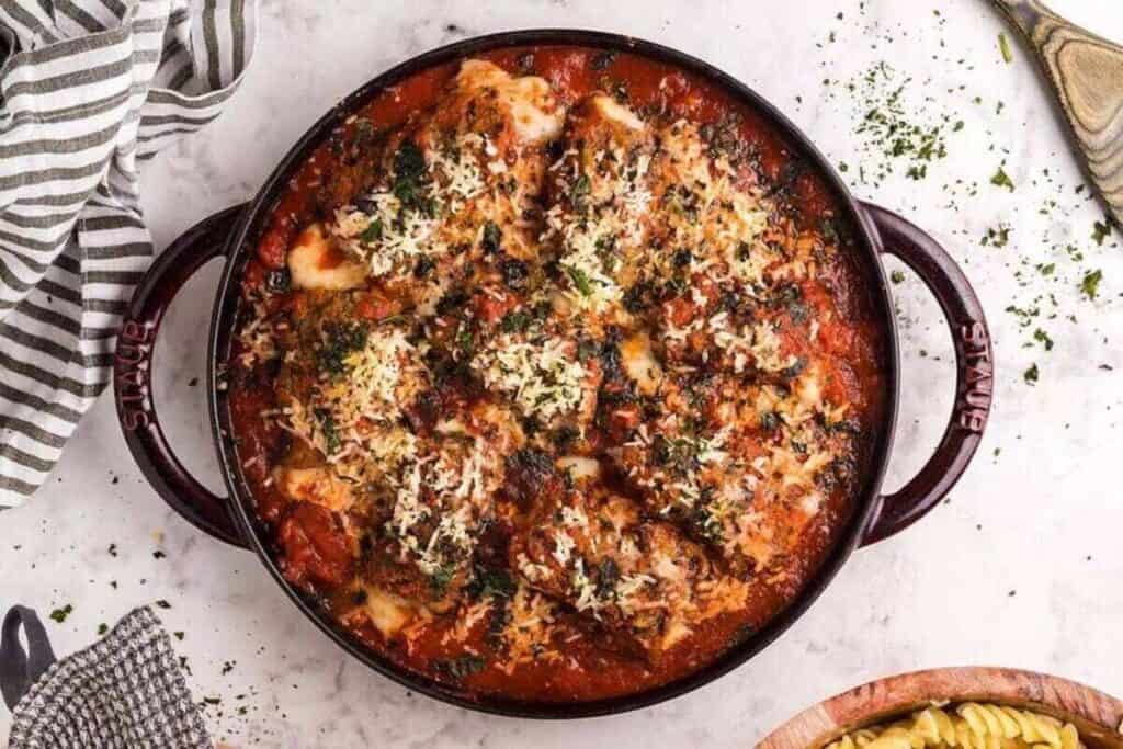 A round baking dish filled with baked chicken parmesan&mdash;perfect for weeknight dinners&mdash;topped with melted cheese and herbs on a marble countertop, next to a striped towel and a bowl of pasta.