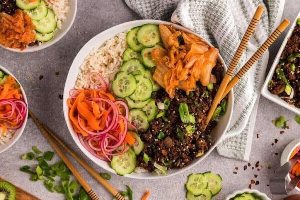 A perfect bowl for Weeknight Dinners, filled with rice, sliced cucumbers, pickled vegetables, kimchi, green onions, and a savory protein, surrounded by small dishes of fresh veggies.