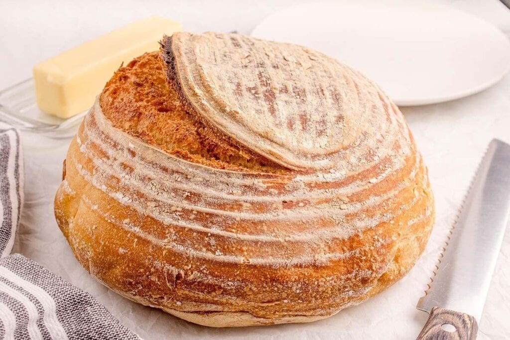 A round loaf of rustic sourdough bread, a classic among baked goods, sits on a white surface near a bread knife, a dish of butter, and a plate.