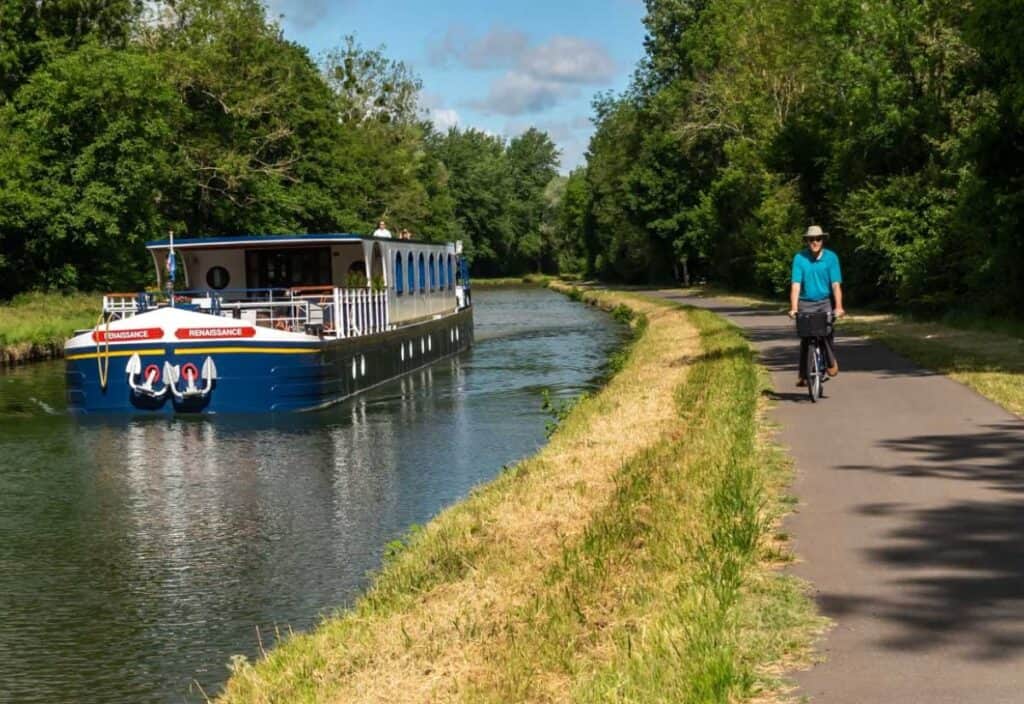 A blue canal boat, offering a luxury barge cruise, travels along a narrow waterway while a man rides a bicycle on the paved path beside the canal, surrounded by greenery.