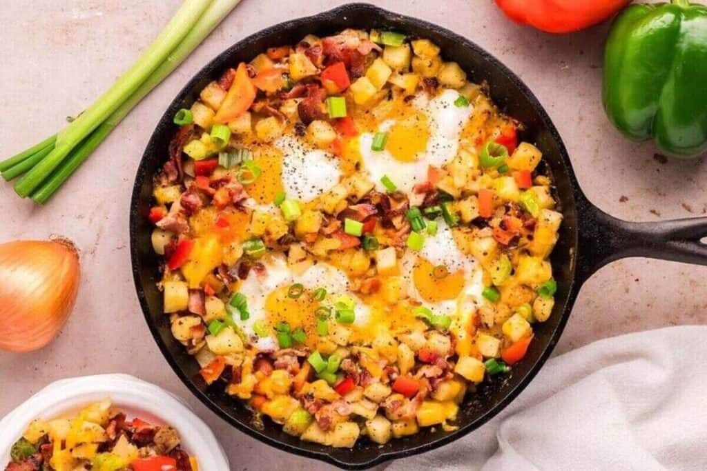 A cast iron skillet filled with breakfast hash made of diced potatoes, bell peppers, eggs, green onions, and bacon&mdash;one of the tastiest breakfast ideas for your morning routine.