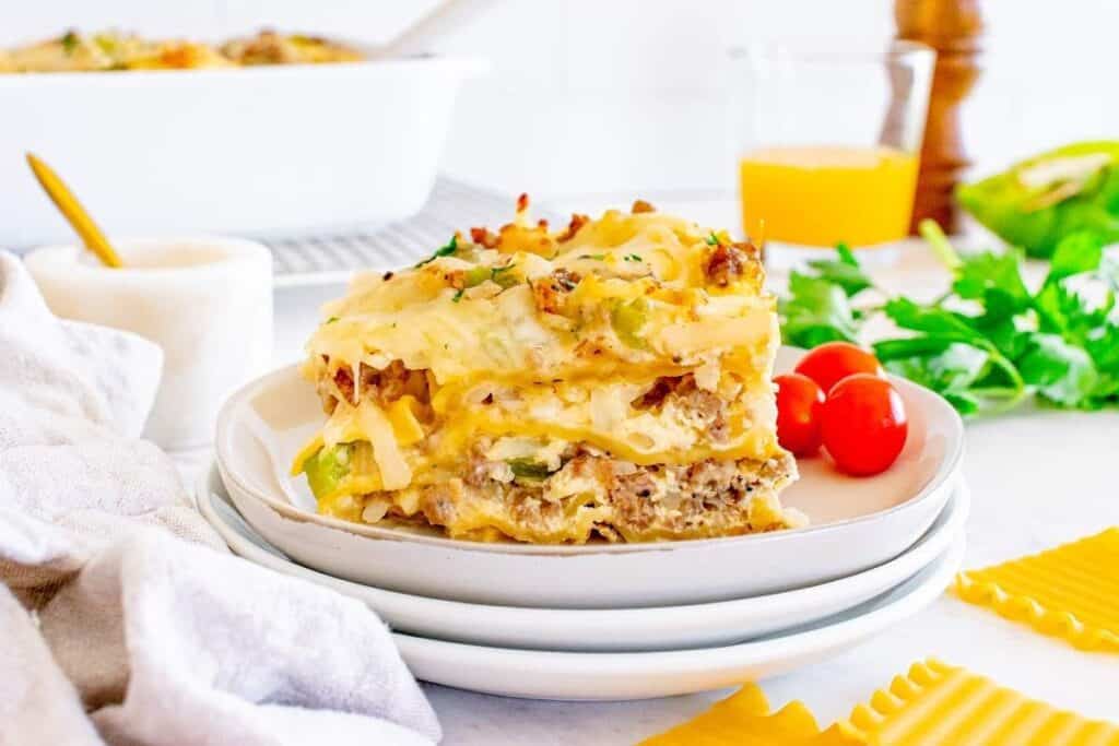 A slice of lasagna with visible layers of pasta, meat, cheese, and vegetables is served on a white plate with cherry tomatoes and herbs&mdash;perfect for those seeking tasty breakfast ideas with a savory twist.