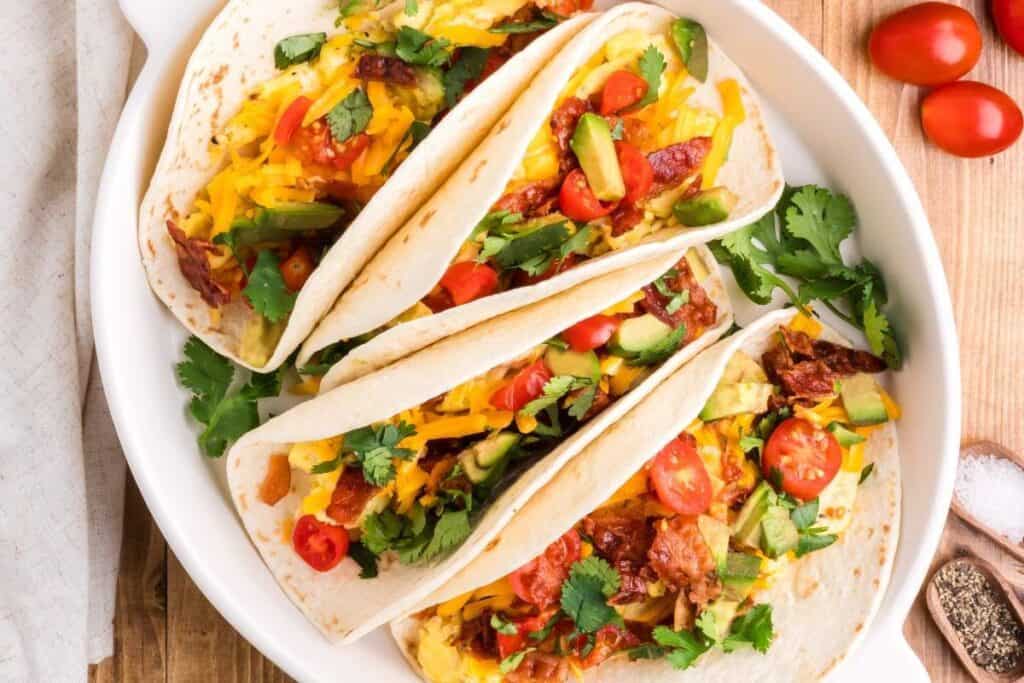 Three soft tacos on a white plate filled with shredded cheese, avocado, cherry tomatoes, bacon, and cilantro&mdash;perfect for those seeking tasty breakfast ideas.