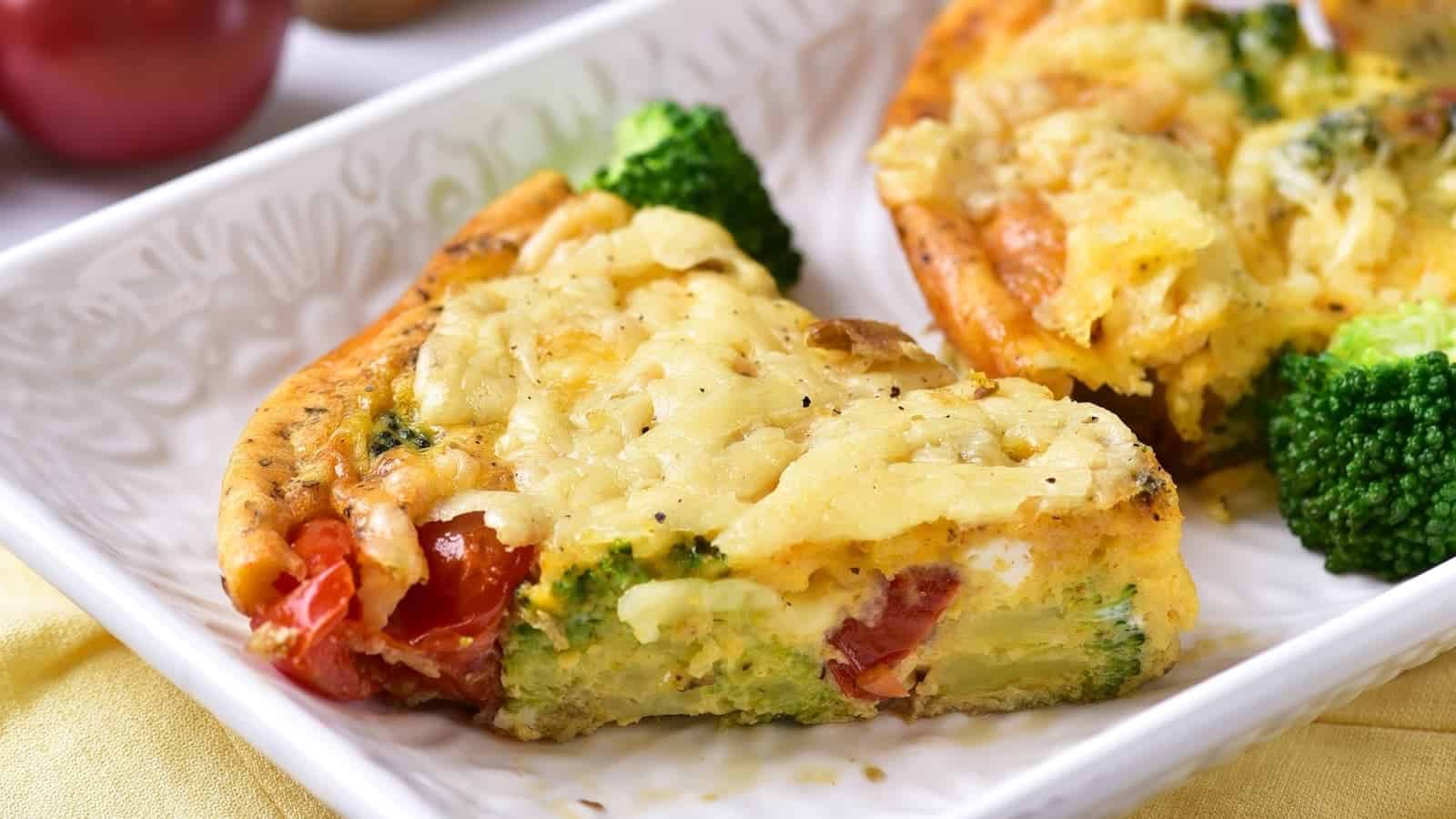 A slice of vegetable frittata with visible broccoli and tomatoes sits on a white plate.