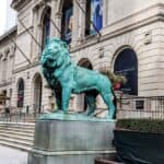 Bronze lion statue stands in front of the Art Institute of Chicago building, with steps and city buildings visible in the background.