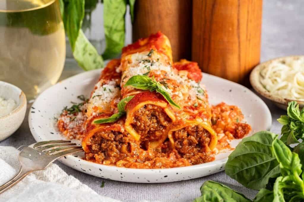 Two cannelloni pasta tubes filled with meat sauce, topped with tomato sauce, grated cheese, and basil—one of our Pasta Classics—served on a white plate with a fork.