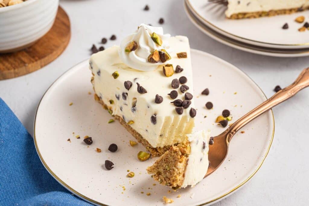 A slice of cheesecake with chocolate chips and chopped pistachios on a plate, topped with whipped cream, with a fork holding a bite-sized piece&mdash;one of those easy desserts that everyone will love.