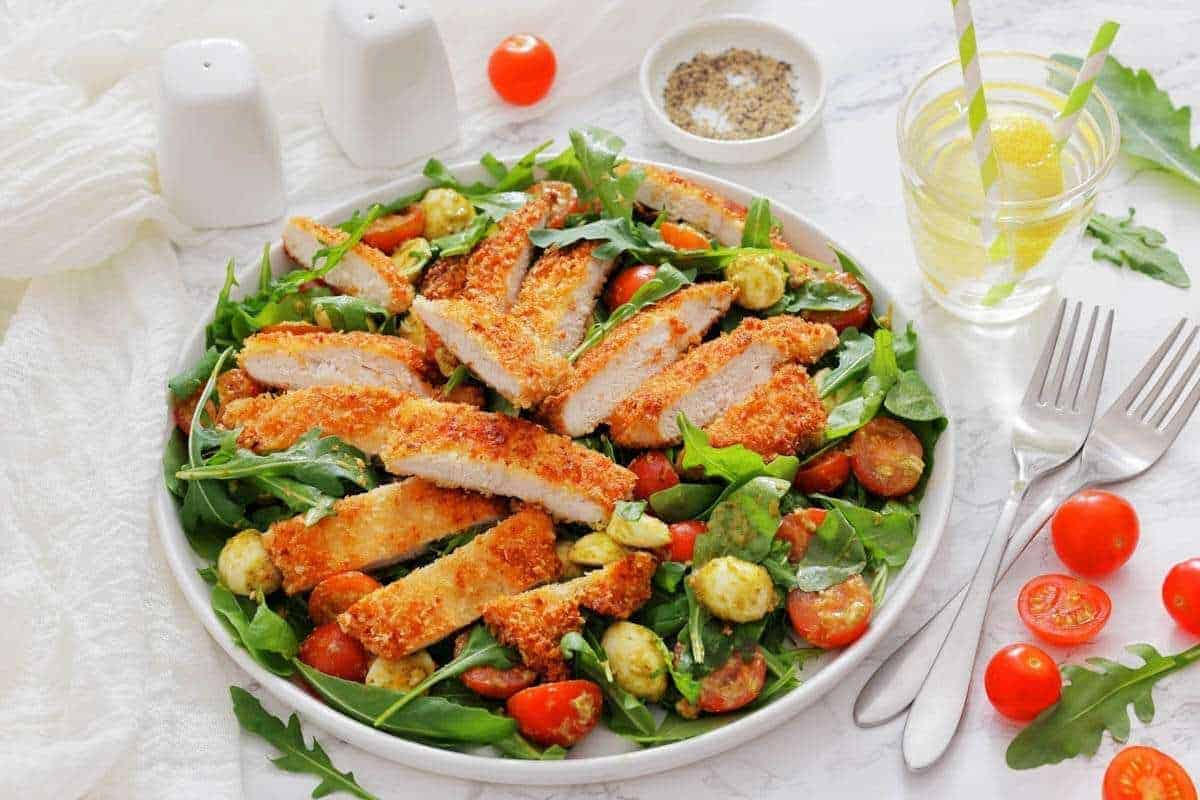 A plate of Chicken Parmesan Salad with a glass of lemon water and utensils on the side.