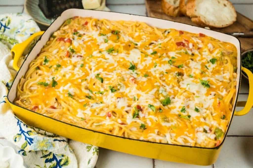 A rectangular yellow baking dish filled with Pasta Classics baked spaghetti, topped with melted cheese and garnished with chopped herbs; sliced bread is in the background.