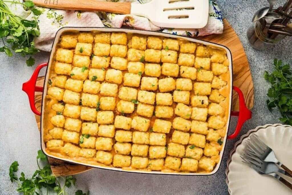 A casserole dish filled with rows of golden tater tots atop a savory baked filling, resting on a wooden board with plates, utensils, and herbs nearby&mdash;perfect for fans of classic casserole recipes.