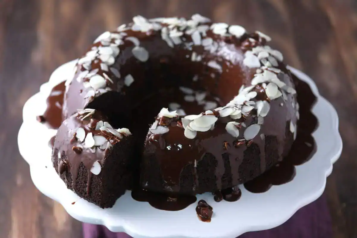 A chocolate bundt cake with chocolate glaze and sliced almonds on top, displayed on a white cake stand with one slice cut.