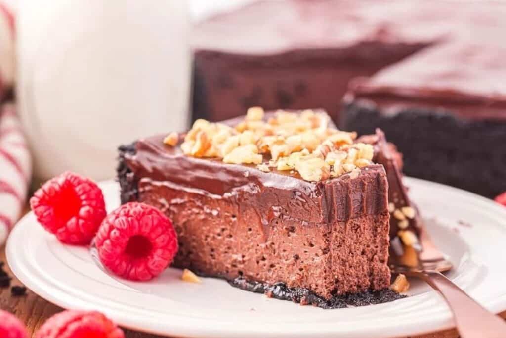A slice of chocolate cheesecake with a chocolate crust, topped with chopped nuts, sits on a plate with fresh raspberries and a fork.