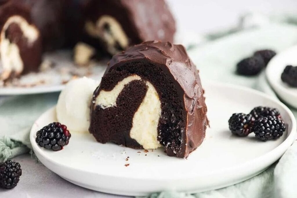 A slice of chocolate bundt cake with a cream cheese filling, served with a scoop of vanilla ice cream and fresh blackberries on a white plate.