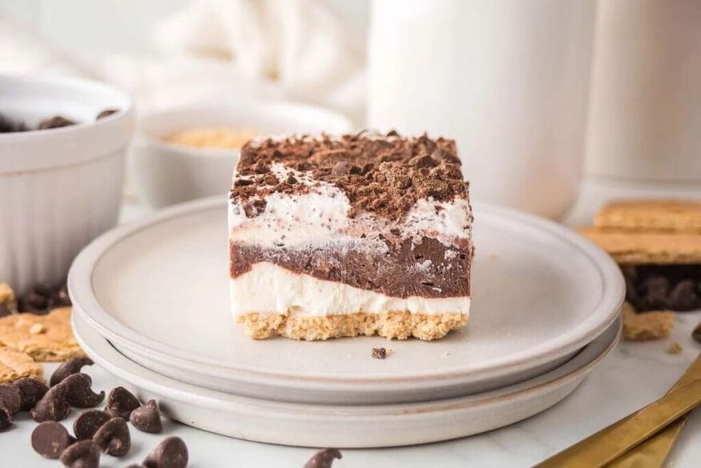A layered dessert bar with a crumbly base, creamy middle, and chocolate topping sits on stacked white plates with chocolate chips and graham crackers nearby.