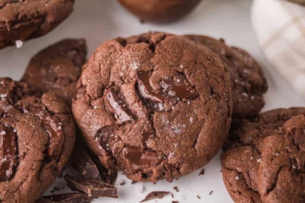 Close-up of several chocolate cookies with chunks of melted chocolate and a light sprinkling of salt on a white surface.