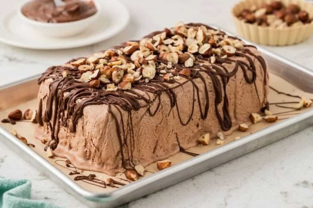 A rectangular chocolate ice cream dessert topped with chopped nuts and drizzled chocolate sits on a metal tray.