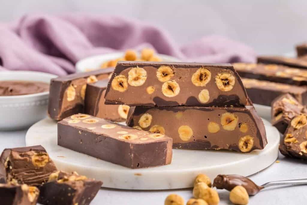 Rectangular pieces of chocolate with whole hazelnuts inside are arranged on a round marble board, with more hazelnuts and chocolate spread nearby.