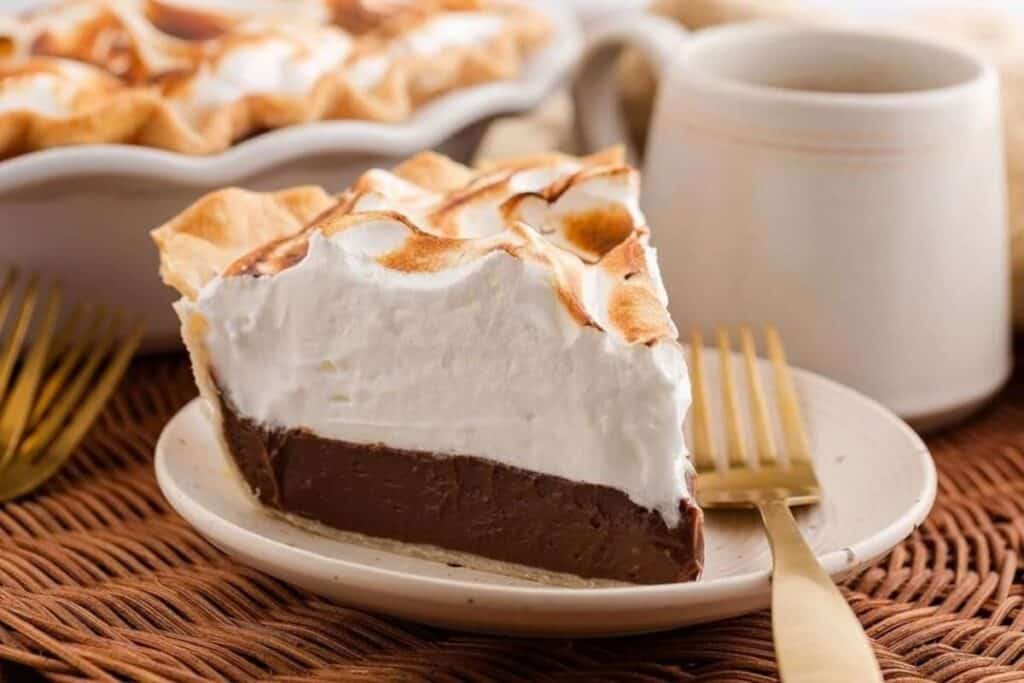 A slice of chocolate meringue pie on a plate with a fork, with a mug and the rest of the pie in the background.