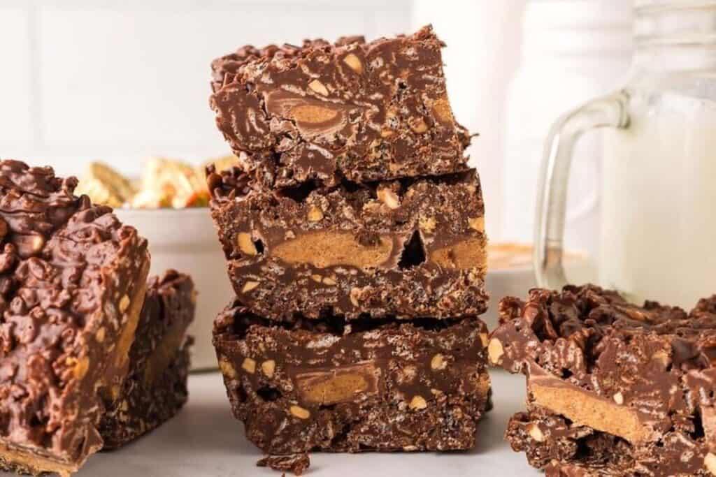 Stacked chocolate peanut butter bars with a crispy texture are displayed on a table next to a glass of milk.