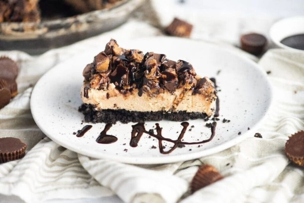 A slice of cheesecake with a chocolate cookie crust, topped with chopped peanut butter cups and drizzled with chocolate sauce on a white plate.