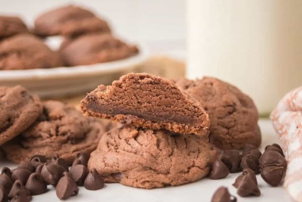 A stack of double chocolate cookies, one split in half to show the gooey chocolate center, with chocolate chips scattered around and more cookies in the background.