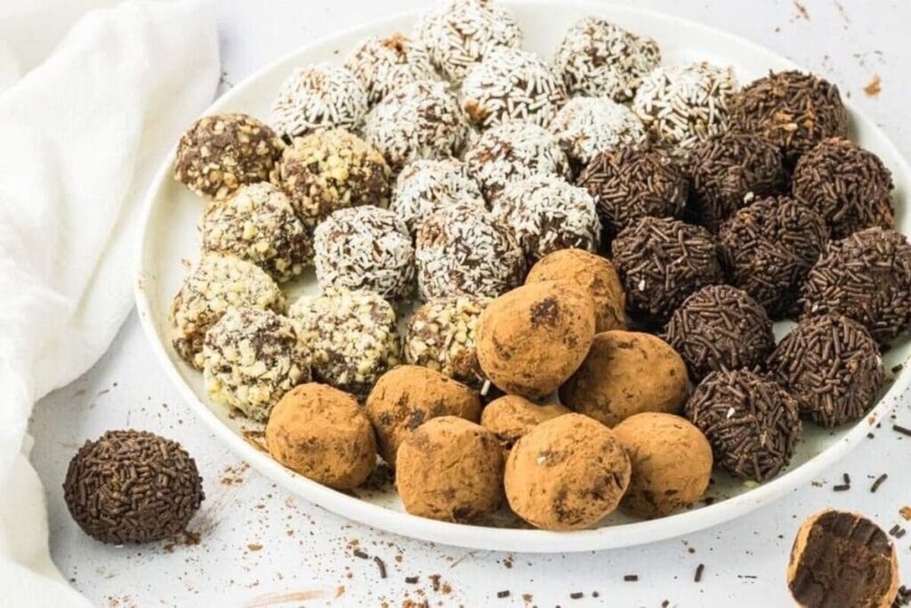 A plate of assorted chocolate truffles coated in nuts, coconut, chocolate sprinkles, and cocoa powder on a white surface.