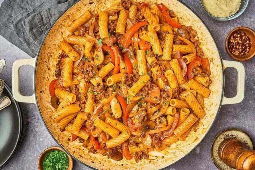 A perfect Weeknight Dinner: a large pan filled with rigatoni pasta, ground meat, sliced red bell peppers, mushrooms, and tomato sauce, surrounded by bowls of grated cheese and red pepper flakes.