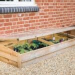 A wooden cold frame with a clear lid contains potted plants and sits on gravel next to a brick house wall and window.