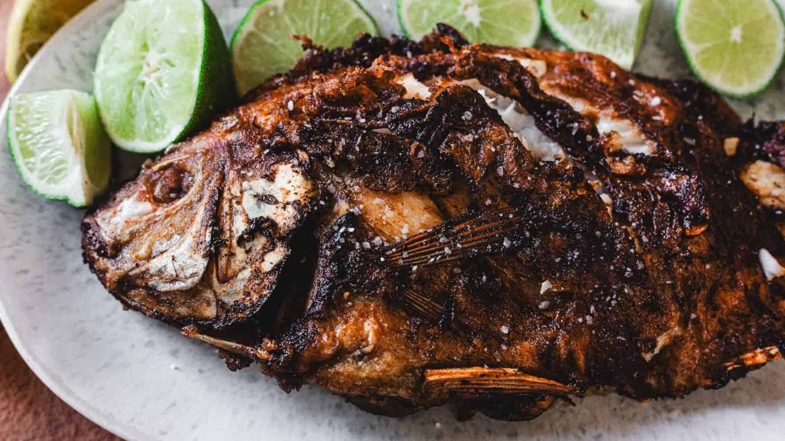 A whole fried fish with crispy, browned skin is served on a plate with several lime wedges arranged around it.