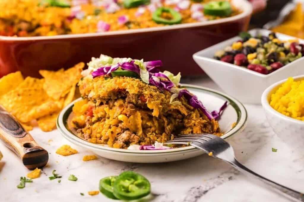 A plate of layered casserole, perfect for Casserole Recipes lovers, is topped with lettuce and cabbage, served with yellow rice and beans. A baking dish and chips appear in the background, while a fork rests on the plate.
