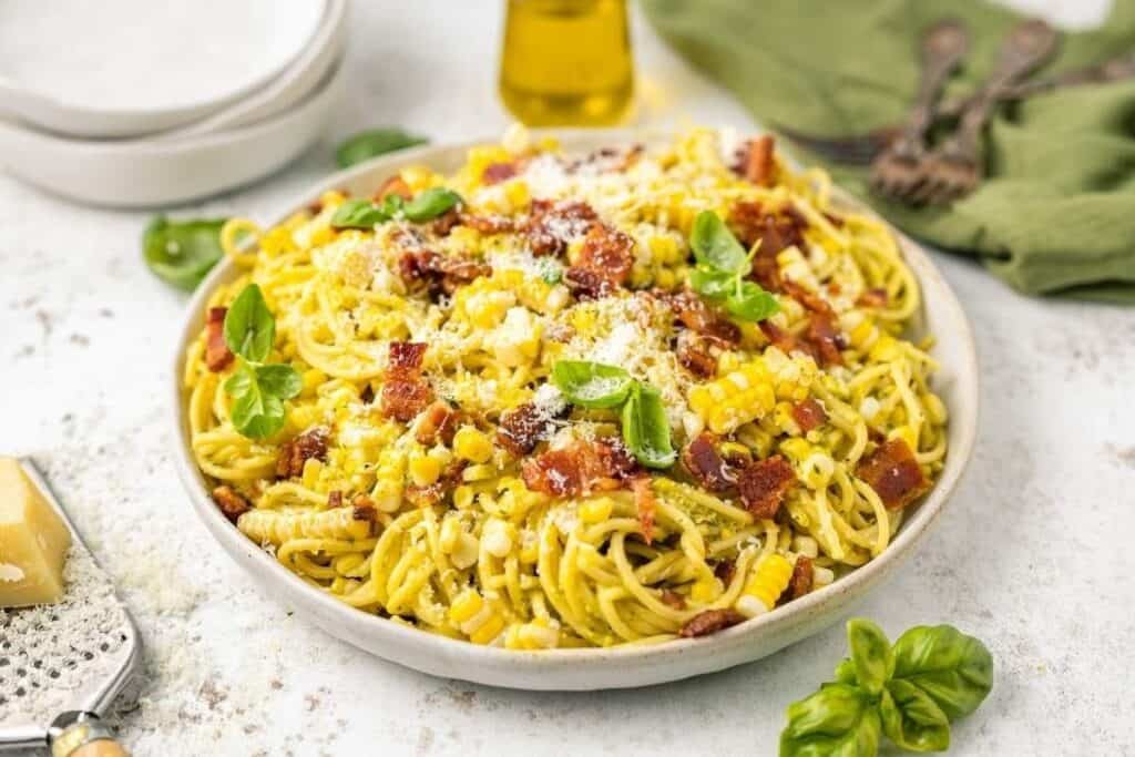A plate of Pasta Classics: spaghetti topped with corn, crispy bacon pieces, grated cheese, and fresh basil, with a grater, basil leaves, and utensils in the background.