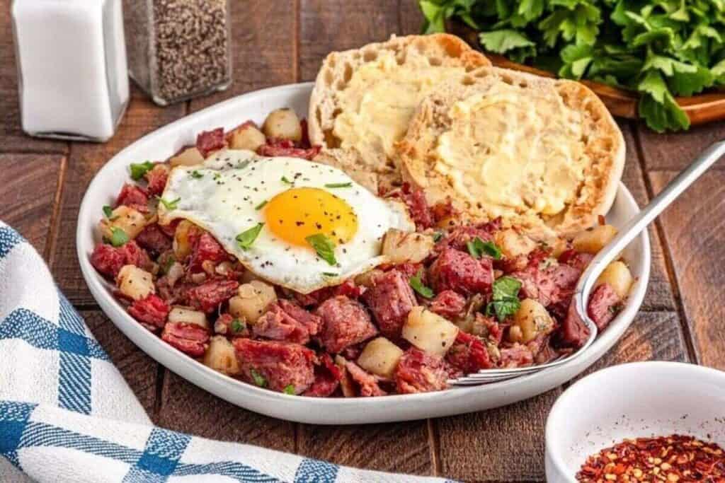 A plate of corned beef hash with diced potatoes, topped with a sunny-side-up egg, and served with buttered English muffins&mdash;perfect if you're searching for tasty breakfast ideas.