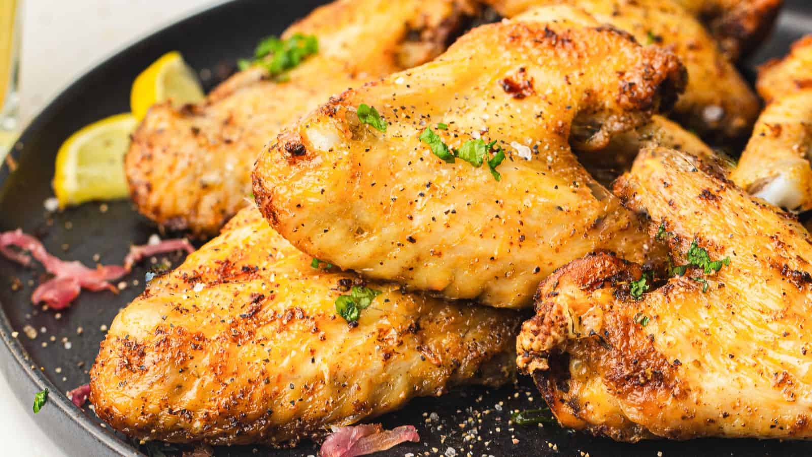 A plate of seasoned and baked chicken wings garnished with chopped herbs, with lemon wedges in the background.