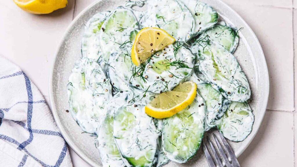 A plate of cucumber slices coated in creamy dressing, garnished with fresh dill, black pepper, and lemon wedges, with a fork on the side.