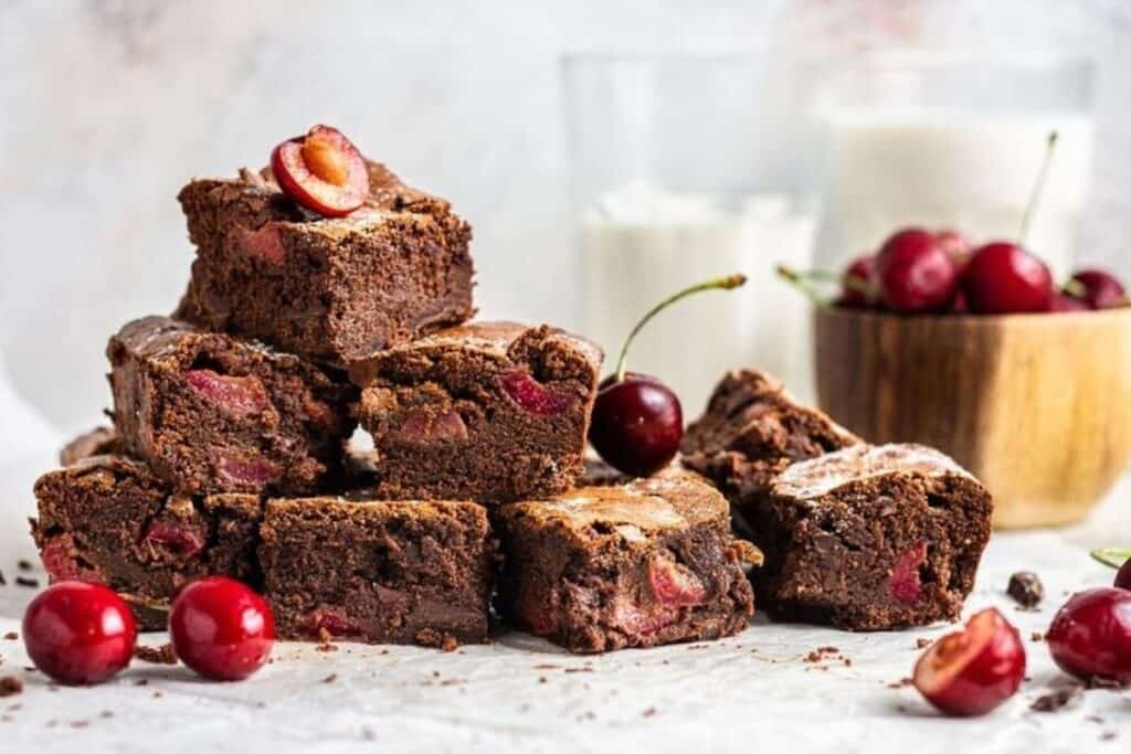 A stack of chocolate brownies with cherries, surrounded by fresh cherries and two glasses of milk in the background.