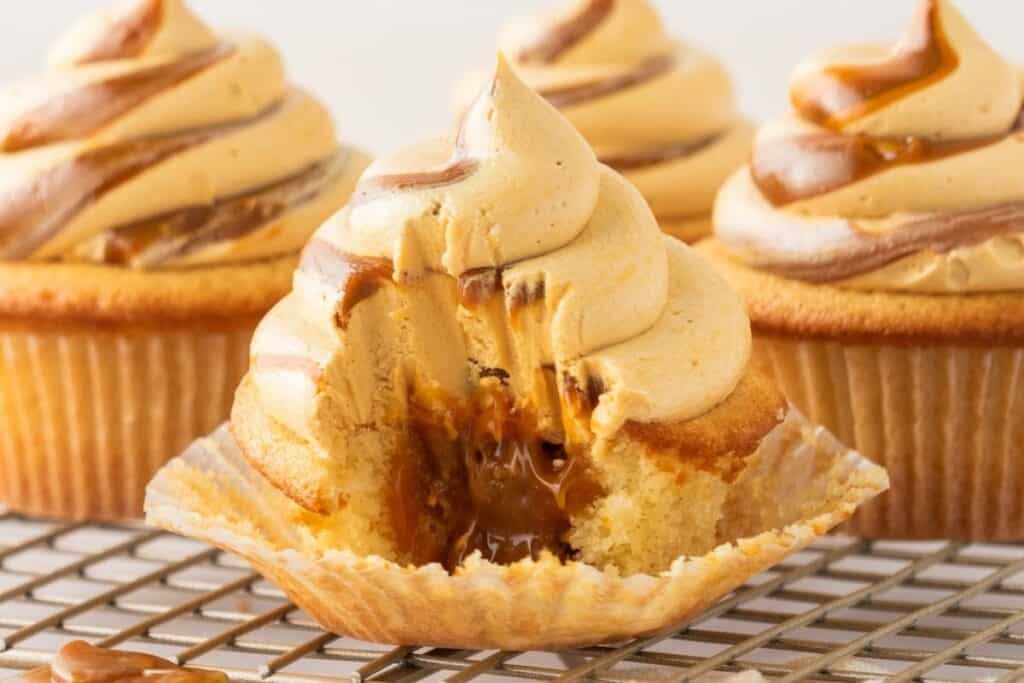 A partially unwrapped cupcake with caramel filling and swirled caramel frosting sits on a cooling rack, surrounded by similar cupcakes&mdash;perfect for those seeking easy desserts that look as delicious as they taste.