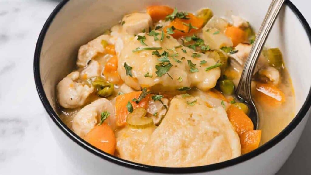 A bowl of chicken and dumplings with carrots, celery, and herbs, served with a spoon.