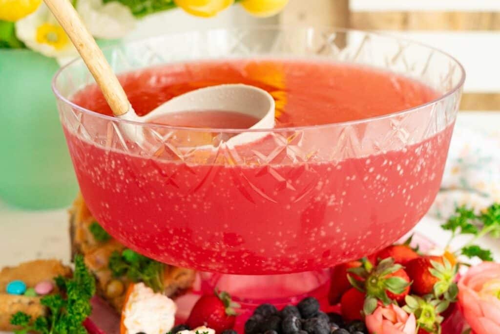 A large glass punch bowl filled with pink bubbly punch and a ladle, surrounded by assorted fruits and snacks&mdash;perfect for serving at gatherings inspired by Bridgerton recipes.