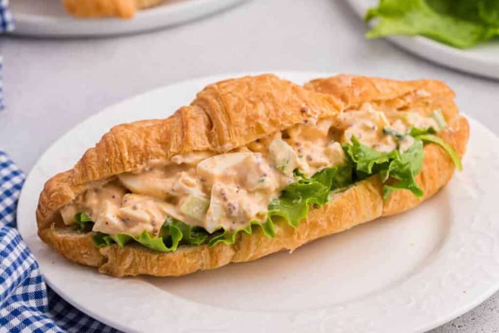 A croissant sandwich filled with lettuce and creamy chicken salad sits on a white plate, perfect for fans seeking Bridgerton recipes to enjoy at an elegant afternoon tea.