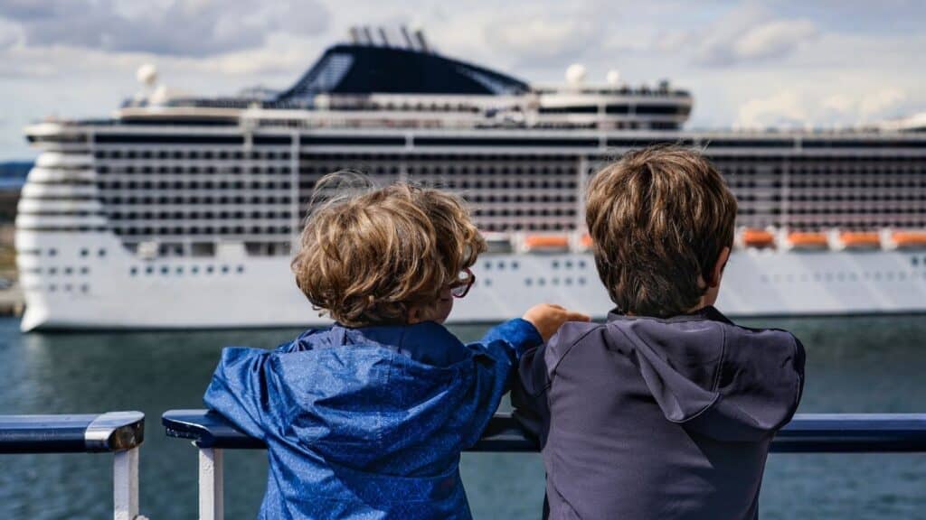 Two children stand on a deck, looking out over the water at a large white cruise ship in the background.