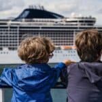 Two children stand on a deck, looking out over the water at a large white cruise ship in the background.