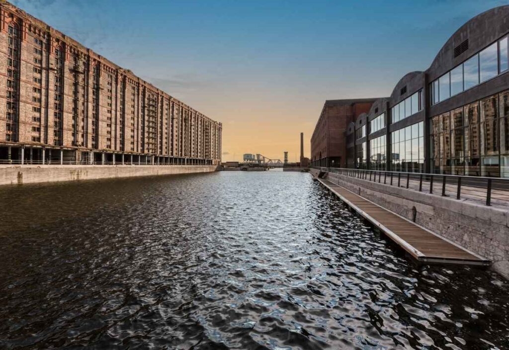 A wide canal flanked by large industrial brick buildings on both sides, with a small dock and railings along the right, under a blue and orange evening sky.