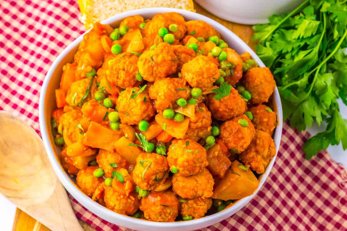 A white bowl filled with a vegetable and meatball stew, garnished with peas and parsley, sits on a red checkered cloth next to fresh parsley, bread, and a wooden spoon.